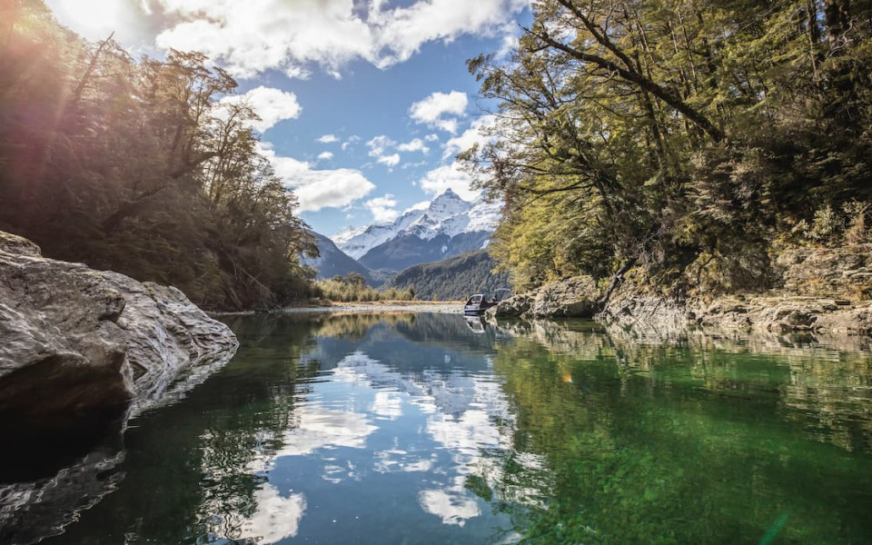 Still water on the Dart River reflecting surrounding trees, with mountains in the distance