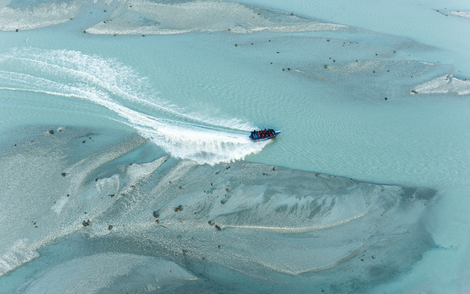 Aerial view of a jet boat speeding along the clear blue Dart River, leaving a spray of water in its wake